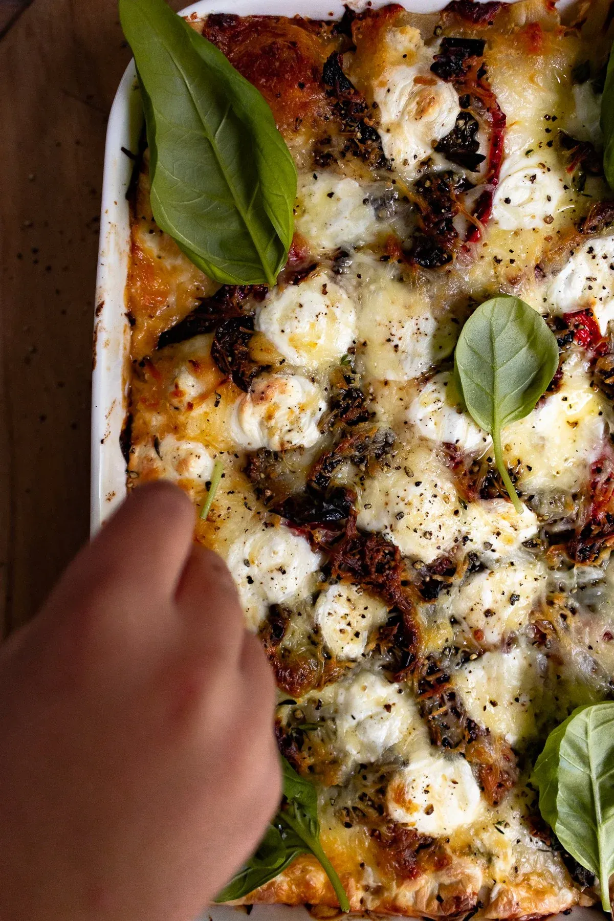 Close angled overhead view of Spicy Chicken and Spinach Lasagna in a white ceramic baking dish with a hand reaching in from lower left