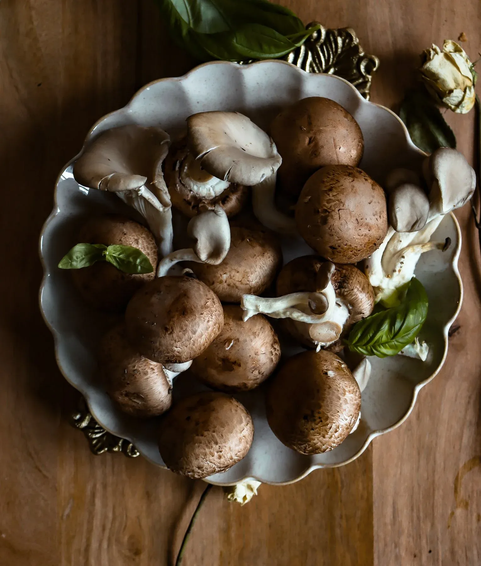 Ingredient prep shot of whole raw cremini and baby bella mushrooms heaped in a white scalloped decorative ceramic bowl on a warm wooden surface