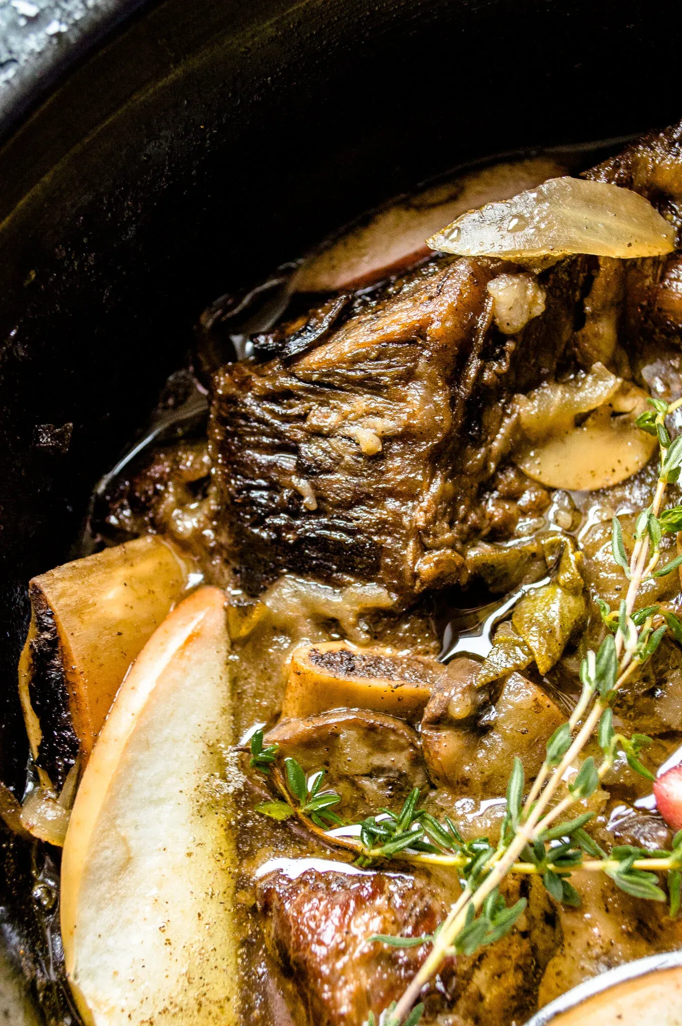 Close-up shot inside a dark slow cooker showing fall-off-the-bone cider-braised short ribs in rich amber braising liquid