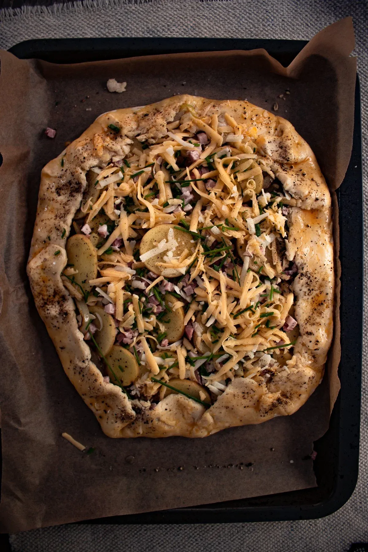 Overhead view of the assembled but unbaked cheesy potato tart on parchment paper in a dark baking pan