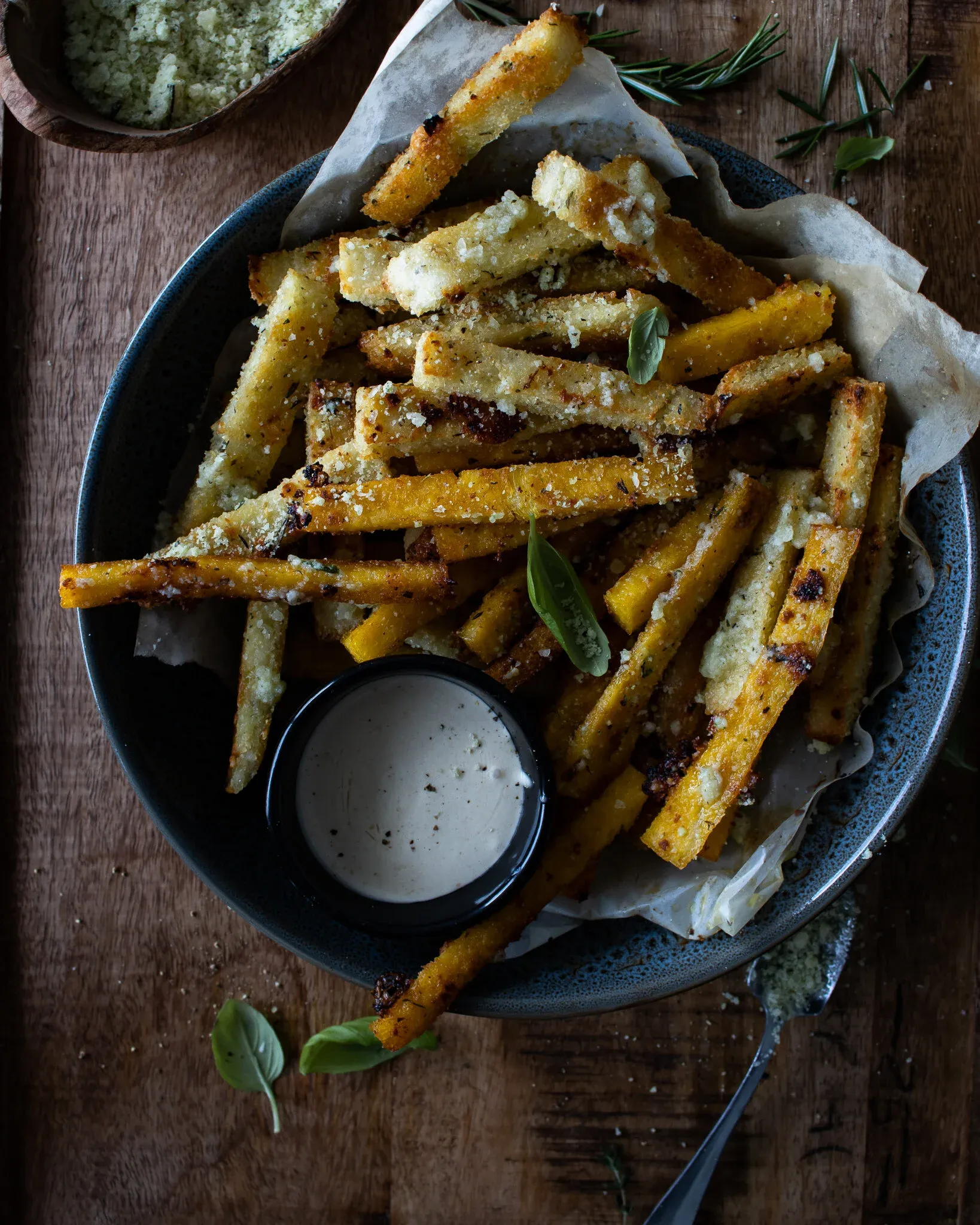 Wider overhead shot of parmesan and herb polenta fries in a dark ceramic bowl on rustic wood