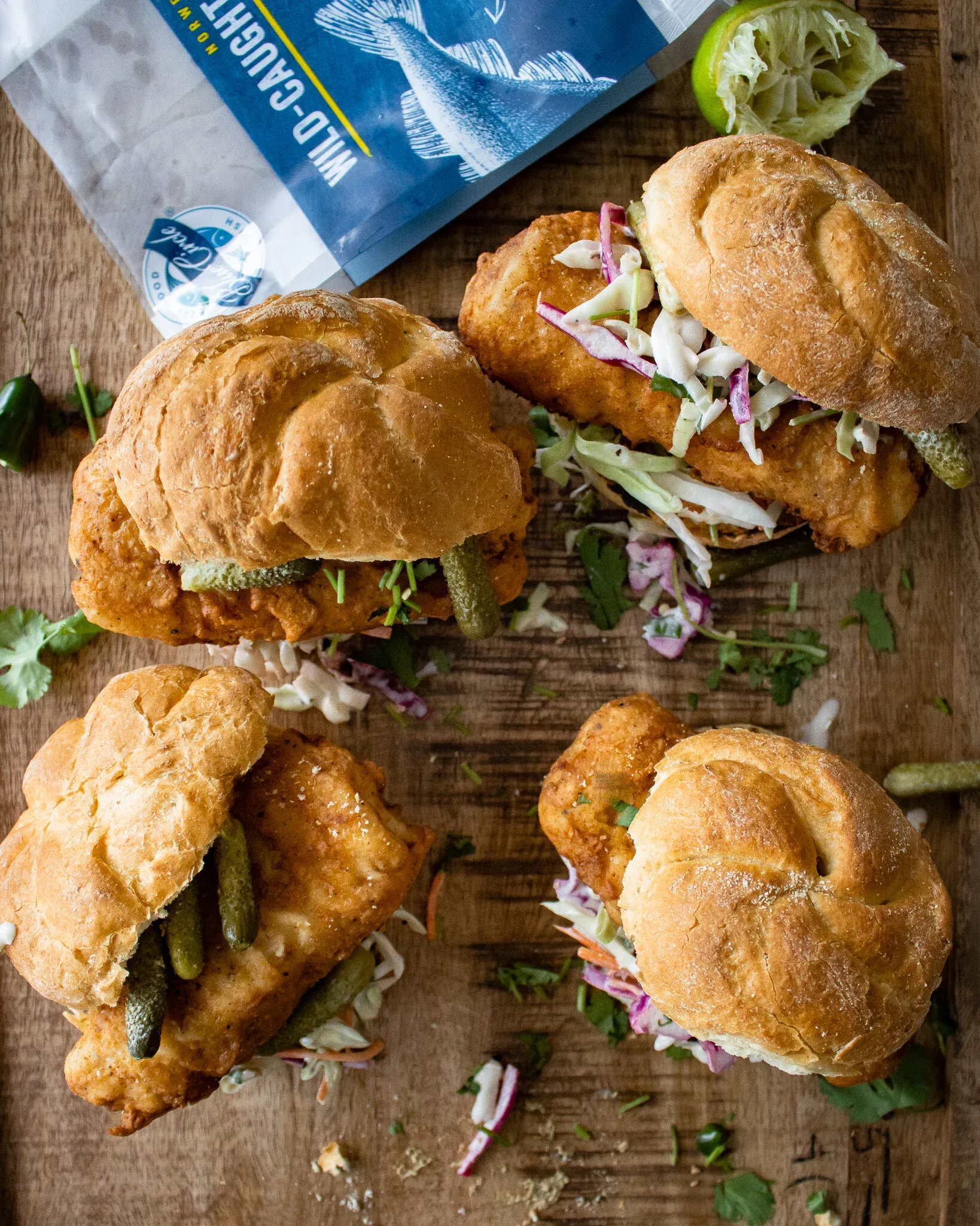 Overhead flat-lay of four crispy fried fish sandwiches on golden crusty rolls arranged on a rustic wooden board