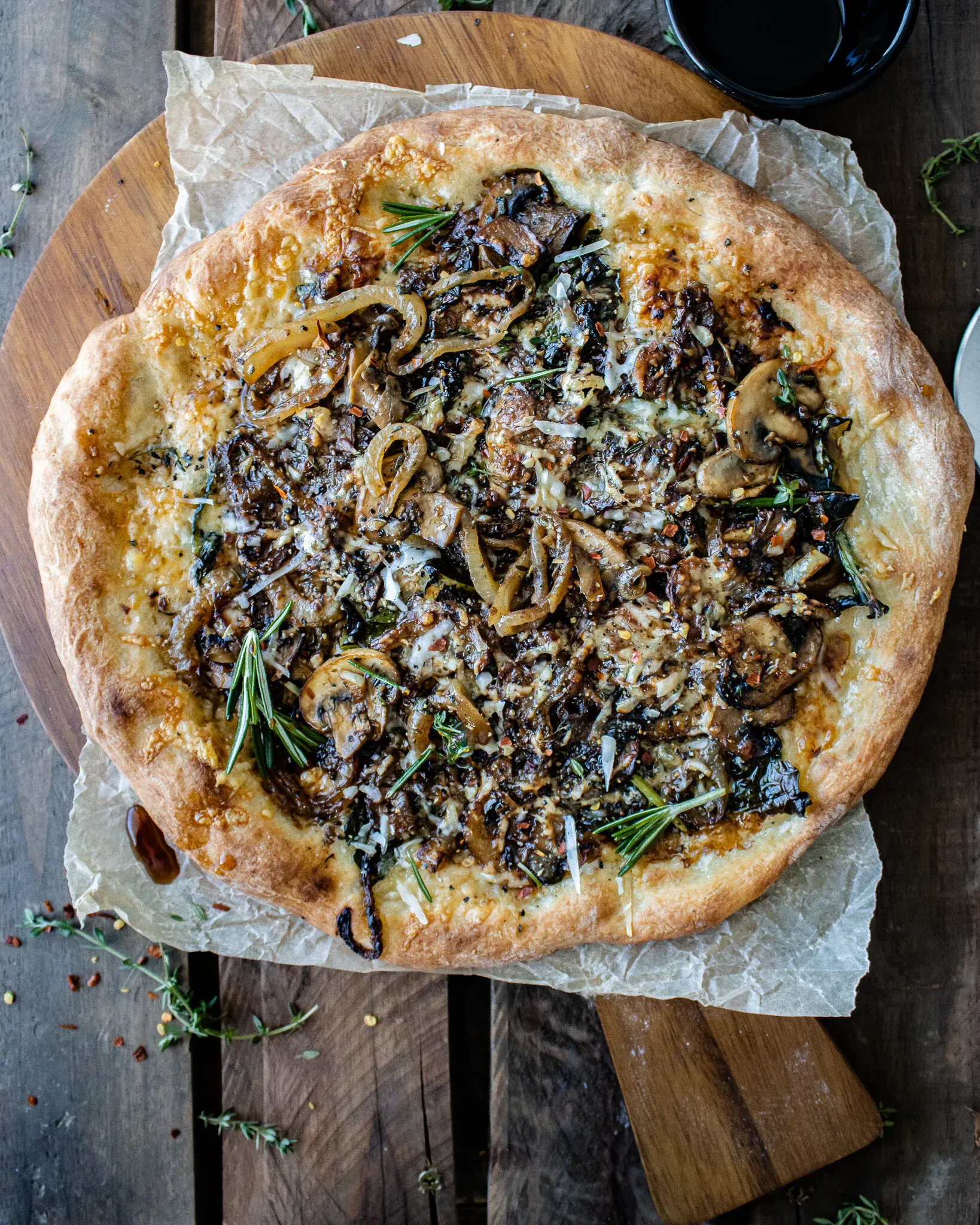 Overhead flat-lay of a whole unsliced caramelized onion and mushroom pizza on a round wooden board over crinkled parchment on a slatted wood surface