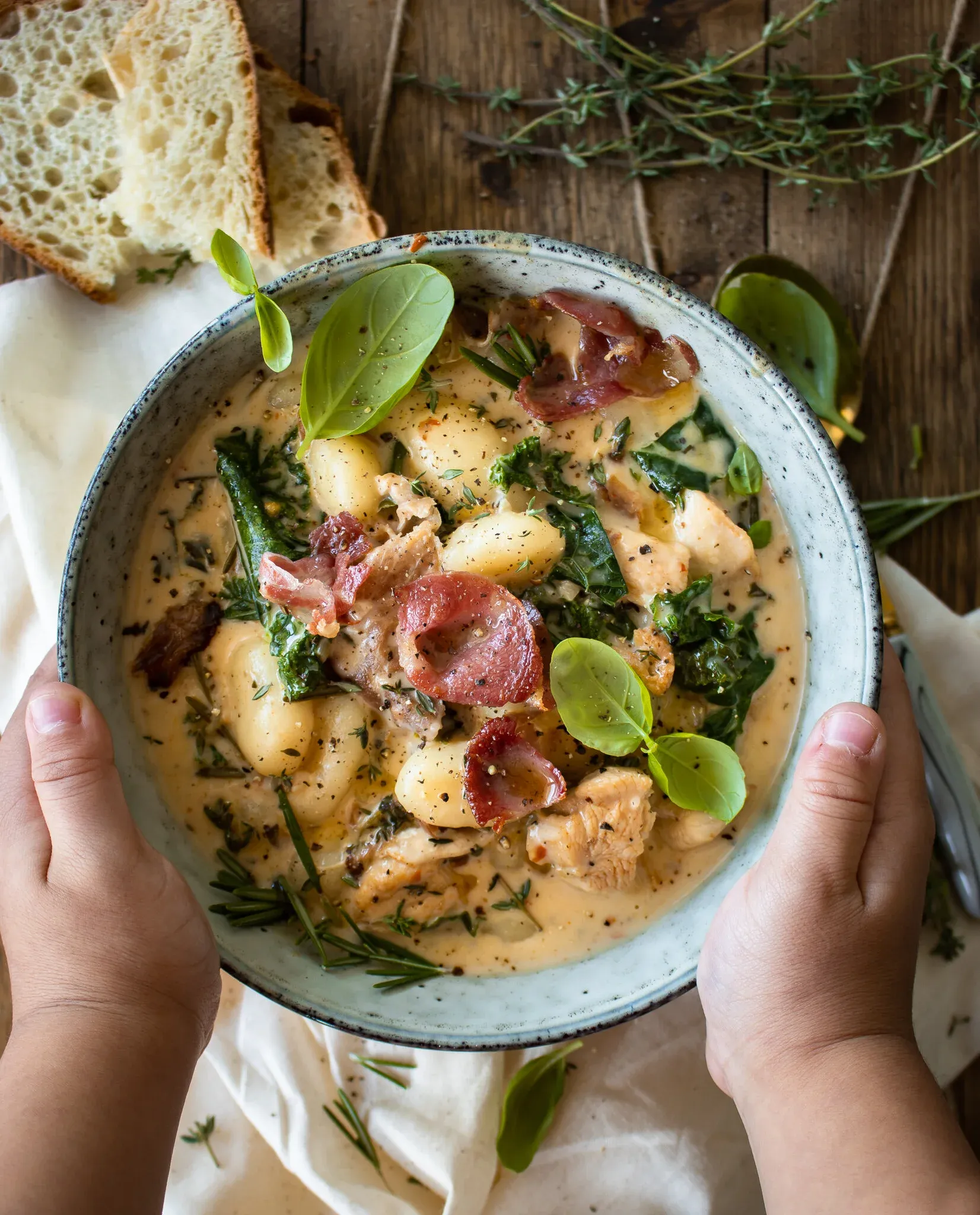 Two hands cradling a gray speckled ceramic bowl of One Pot Gnocchi Soup with Kale and Prosciutto, filled with plump gnocchi
