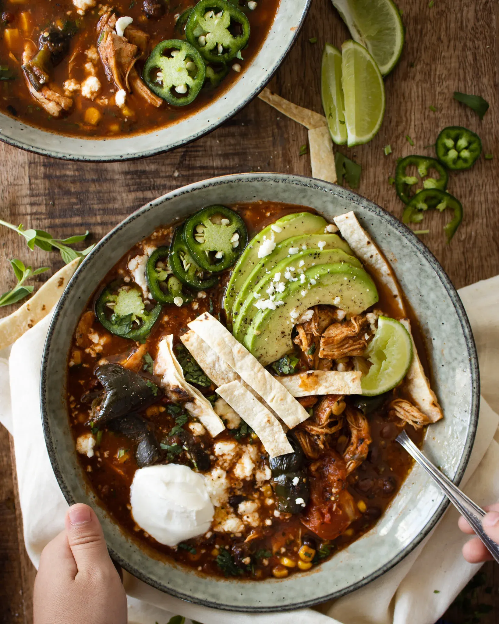 Two hands cradling a ceramic bowl of Chicken Tortilla Soup from opposite sides with a spoon handle visible in the broth