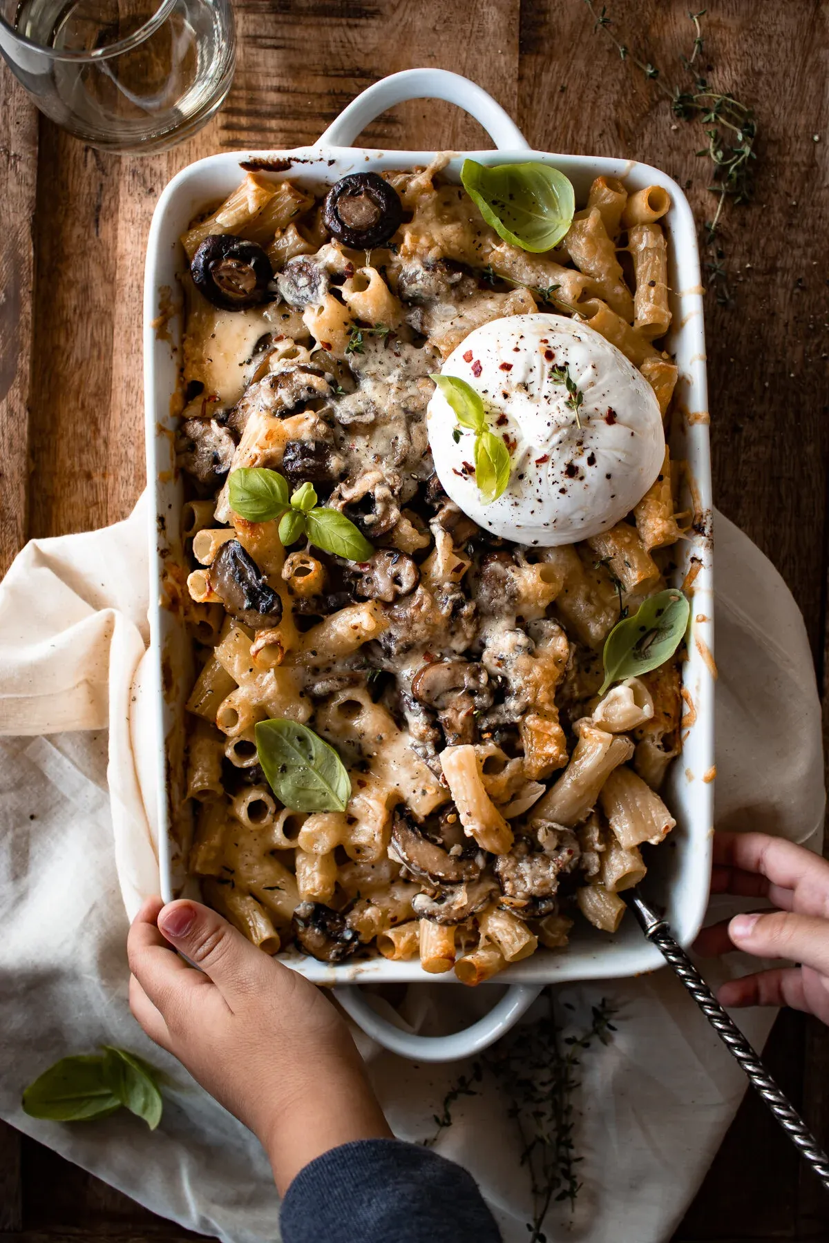 Two hands holding up the white ceramic baking dish of Baked Mushroom Pasta with Burrata over a cream linen napkin on a wood surface