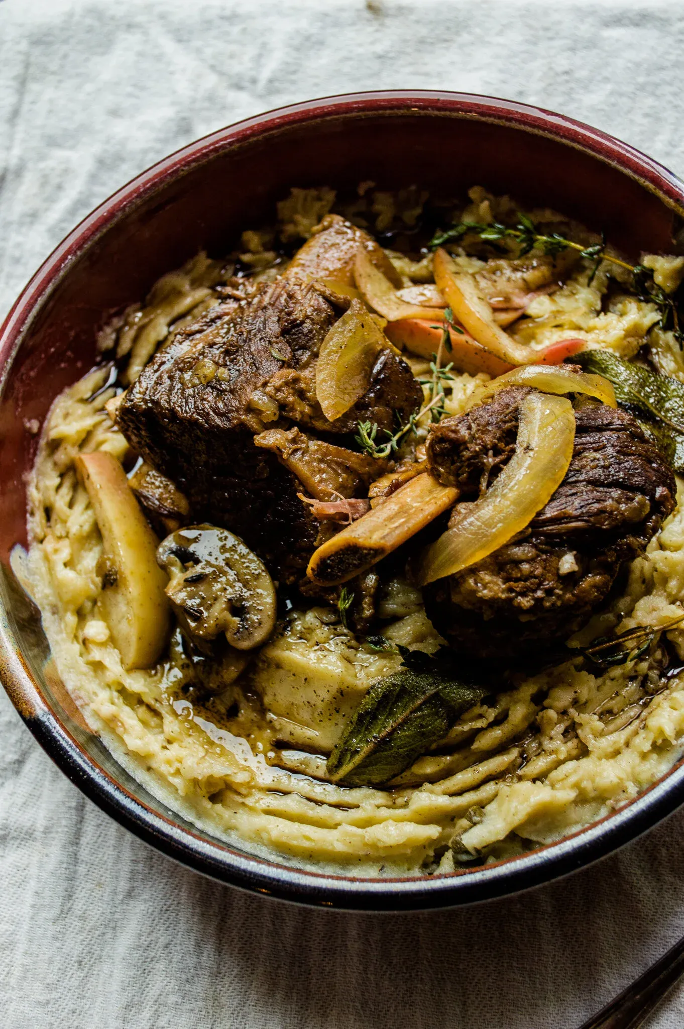 Angled overhead view of slow cooker hard cider braised short ribs with brown butter mashed potatoes in a dark ceramic bowl with red rim on a grey linen cloth