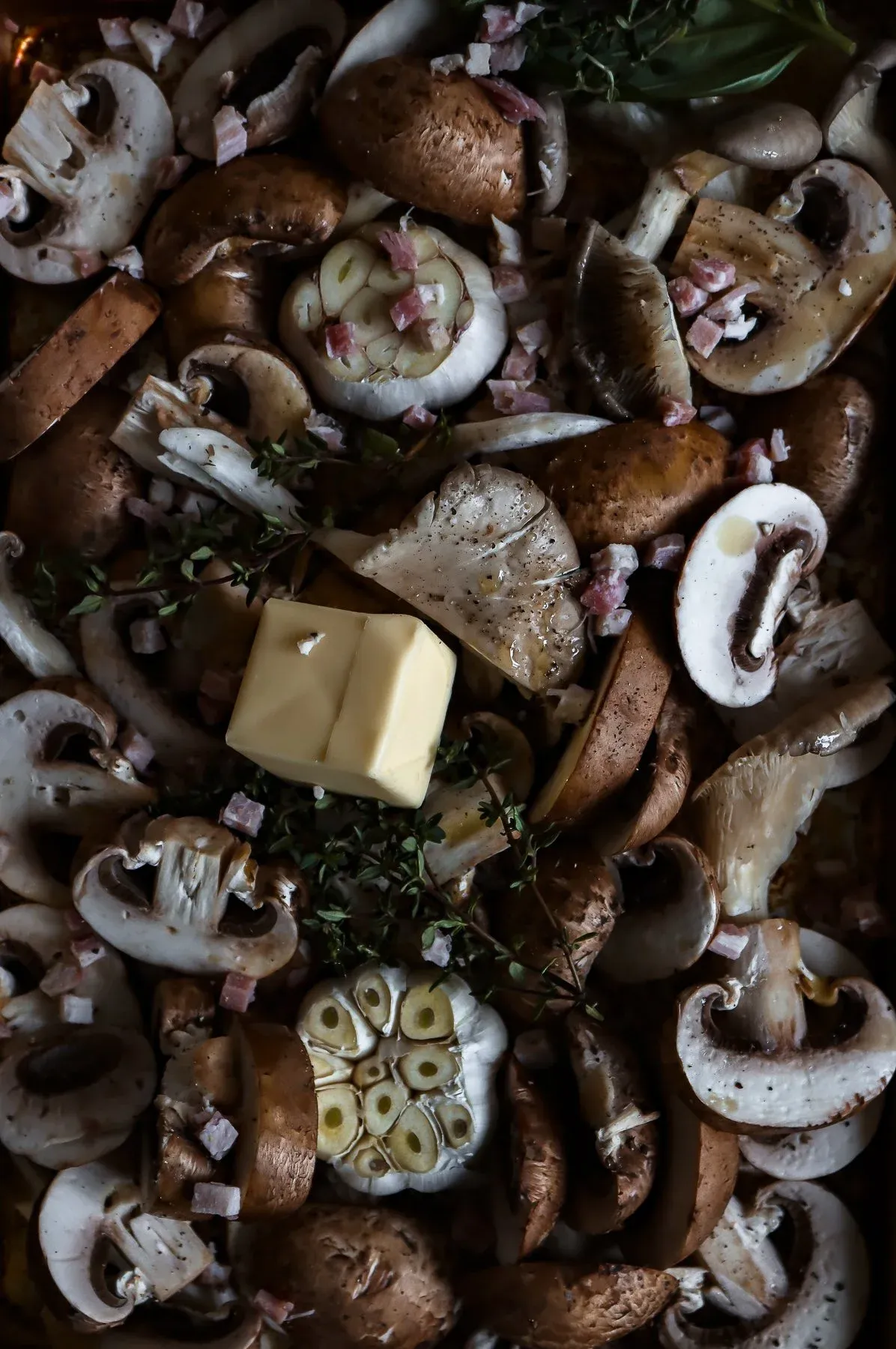 Overhead close-up of sliced and whole cremini mushrooms spread across a dark roasting pan ready to go into the oven