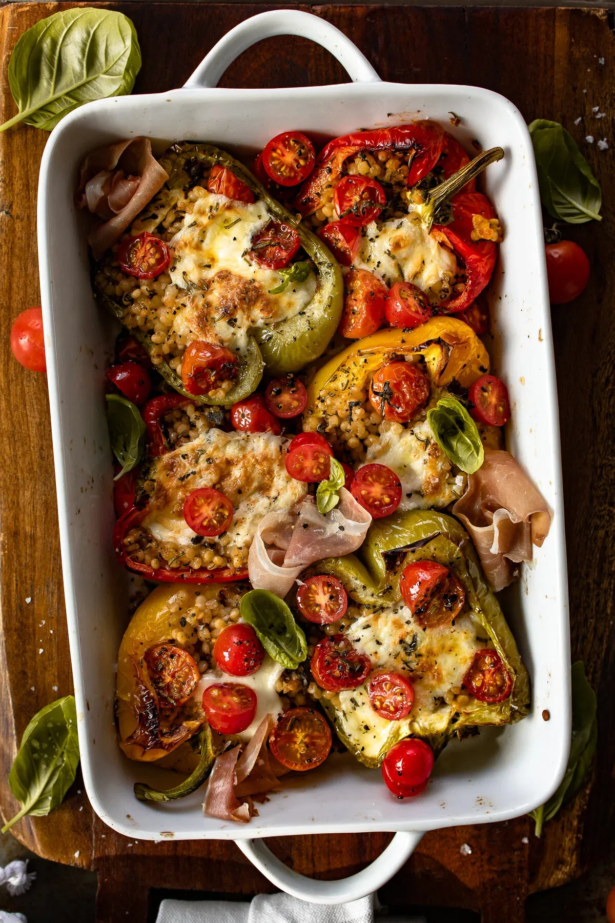 Wide overhead flat-lay of the entire Couscous Stuffed Roasted Bell Peppers with Fresh Mozzarella in the white handled baking dish centered on a dark wood surface