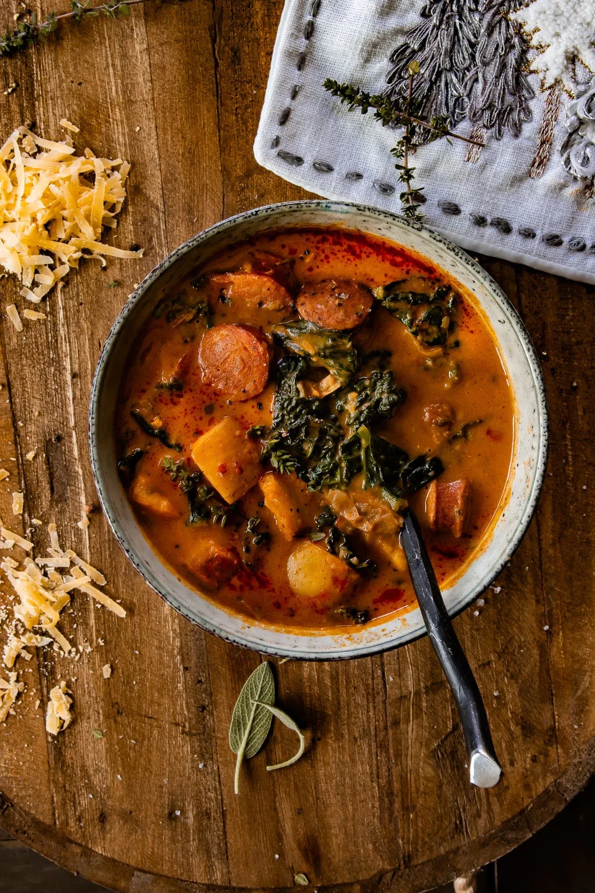 Angled overhead view of Smoked Potato and Sausage Soup in a speckled ceramic bowl on a round wooden surface with a dark spoon