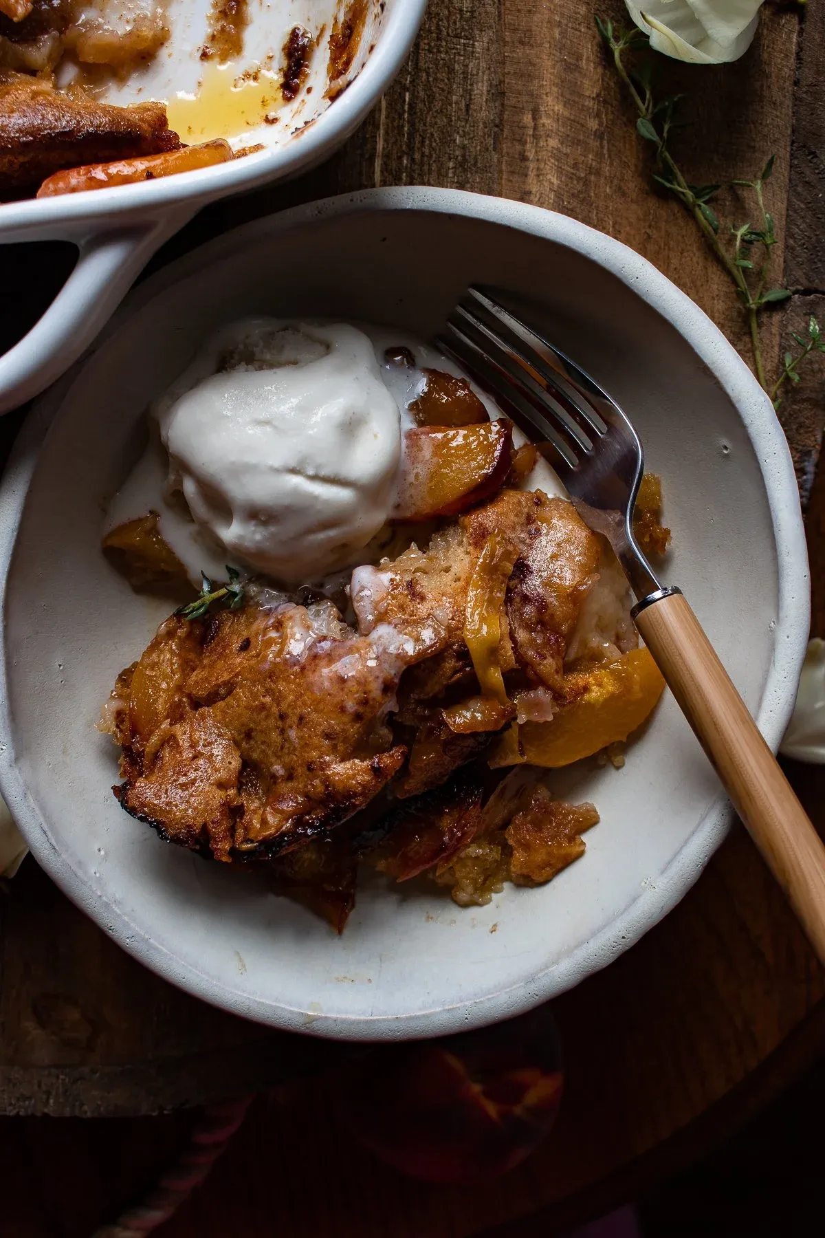 Single serving of Brown Butter Peach Cobbler in a rustic white bowl with one scoop of vanilla ice cream melting alongside golden cobbler chunks and syrupy peach slices