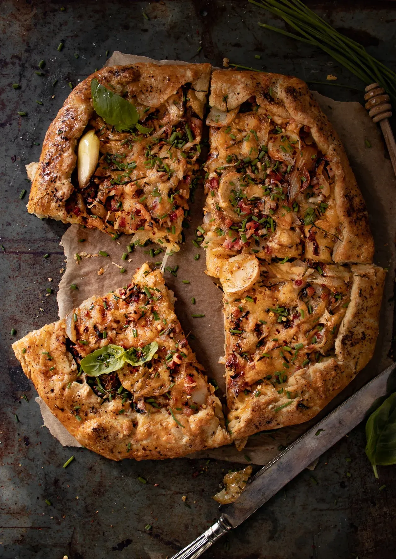 Overhead view of the whole cheesy potato and pancetta galette tart sliced into four wedges on parchment paper on a dark baking sheet