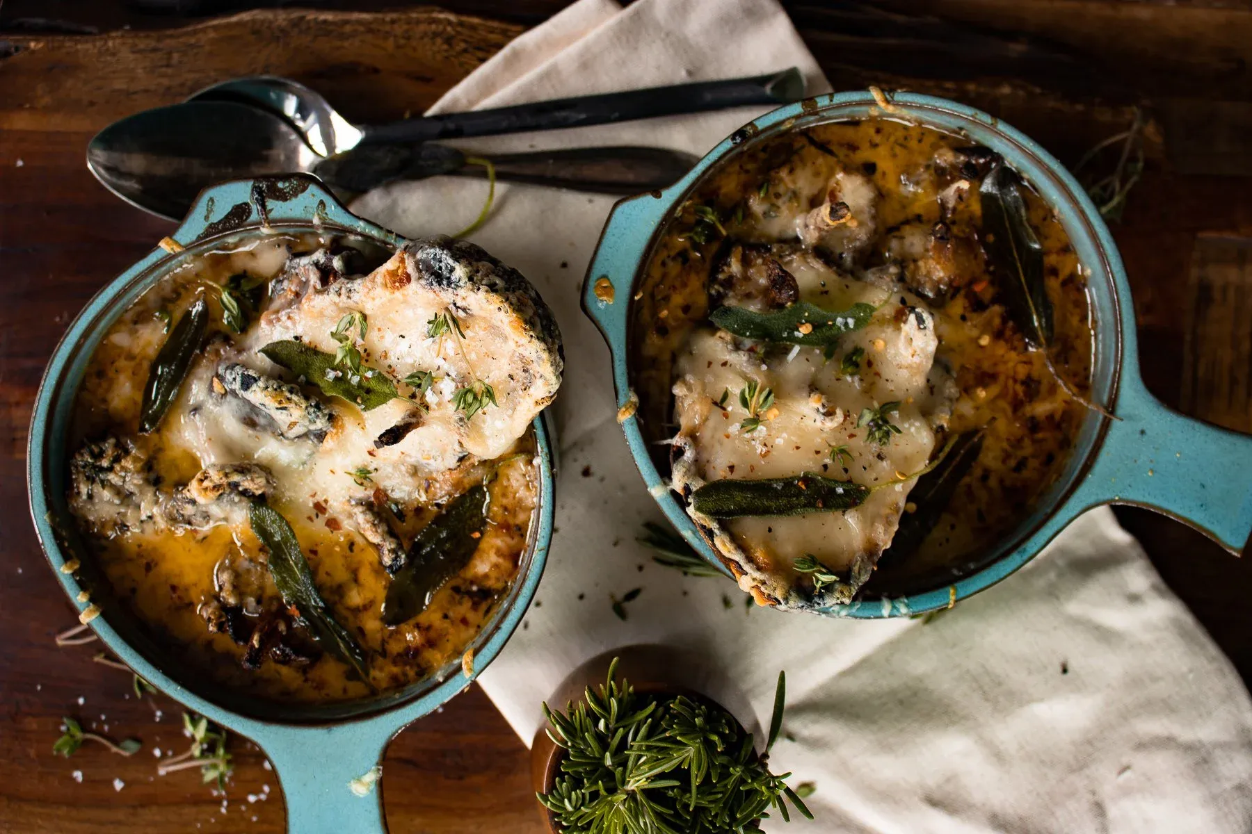 Overhead flat-lay of two teal ceramic crocks of French Onion Soup with Roasted Mushrooms sitting side by side on a dark wood board over a cream linen