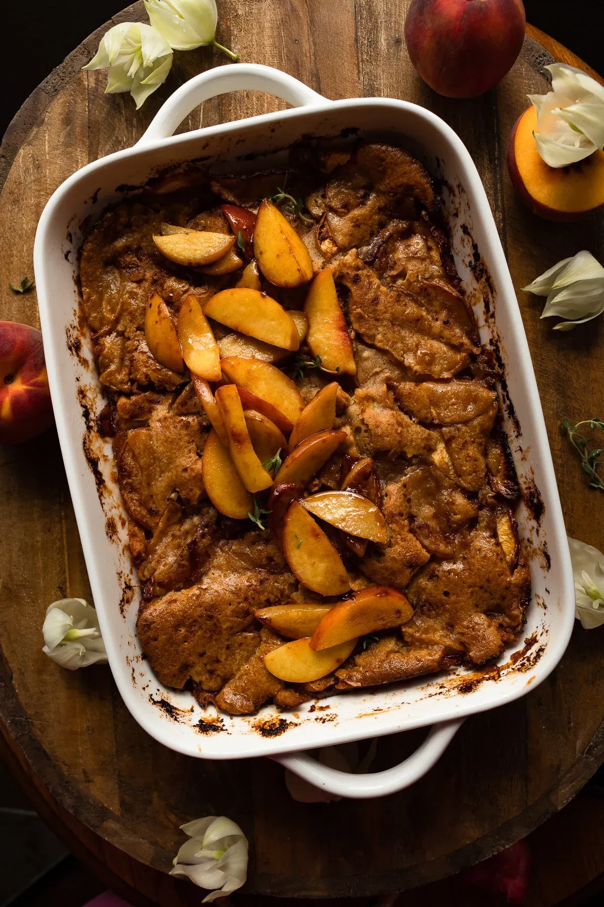 Wider overhead view of Brown Butter Peach Cobbler in the white rectangular baking dish on a dark round wooden surface