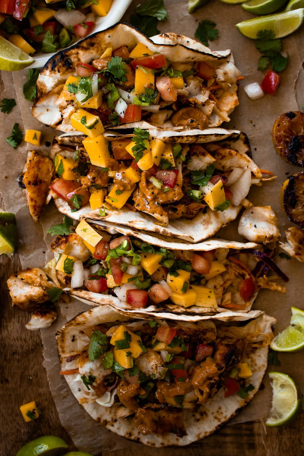 Overhead flat-lay of four Caribbean fish tacos with mango salsa lined up on crinkled parchment paper on a wooden cutting board