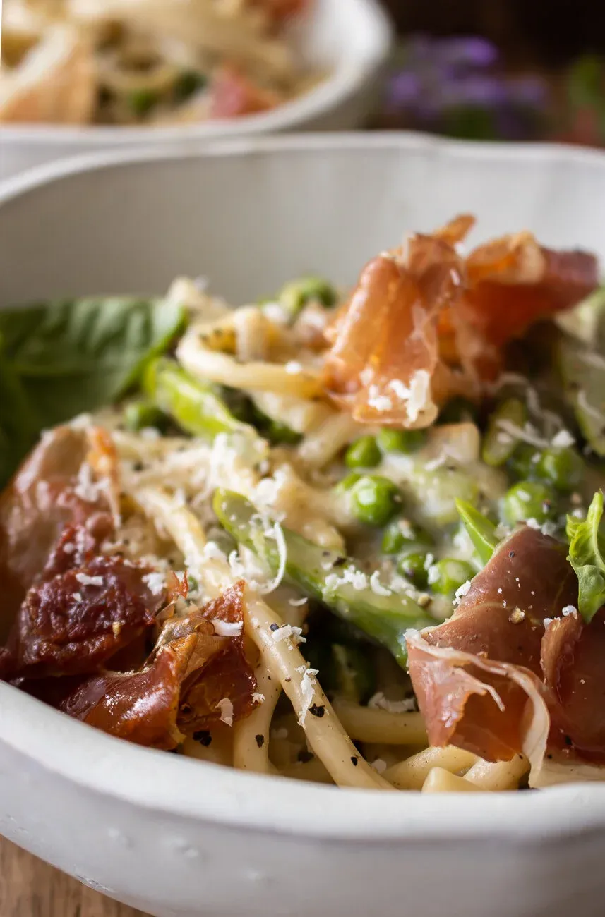 Close-up side-angle of a white bowl of Cacio e Pepe with Spring Veggies and Crispy Prosciutto