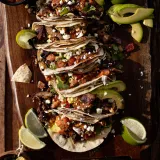Pernil Tacos lined up diagonally on a large dark wooden cutting board