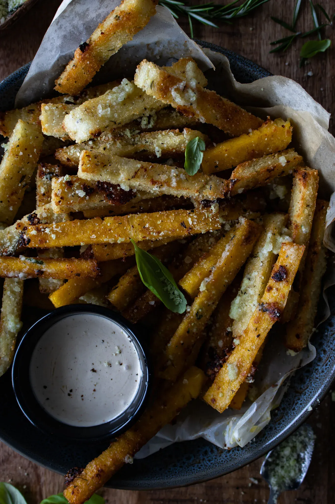 Overhead close-up of parmesan and herb polenta fries piled in a dark navy speckled ceramic bowl lined with parchment paper